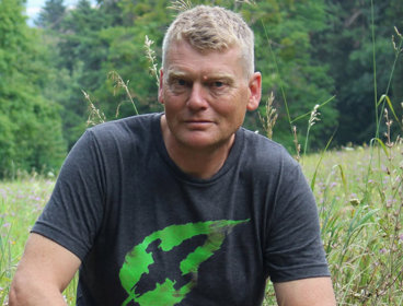 Portrait photograph of Tom Heap in a forest against foreground of flowers and wild grass.