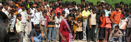 A large group of people gathered around a person with a metal bowl placed on a metal barrel. 