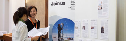 Two people chatting in front of a board of the Royal Geographical Society that says join us. 