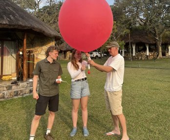 Three people stood on grass in front of a house holding a giant balloon with measuring equipment attached. 