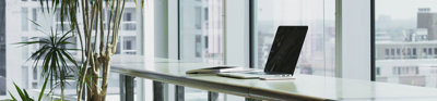 An open laptop on a table next to a wide set of tall glass windows in a modern office.