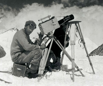 Historic black and white photograph of two explorers taking photographs in a snowy mountainous environment.