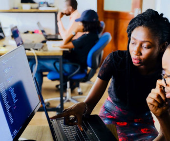 Two people sat at a desk in an office in front of a monitor and a laptop. One person is pointing to the laptop screen. There are other people working at a desk in the background.