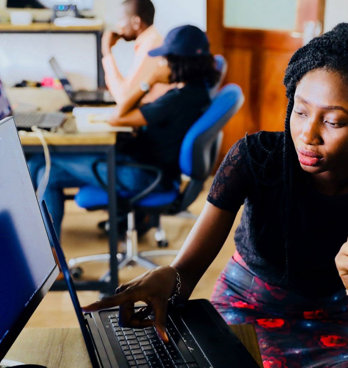 Two people sat at a desk in an office in front of a monitor and a laptop. One person is pointing to the laptop screen. There are other people working at a desk in the background.