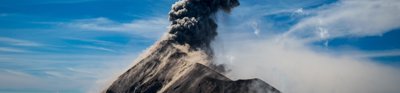 Dense smoke and ash erupt from the mouth of a volcano and ascend into the cloudy blue sky above