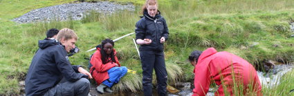 A group of teachers collected by a small fast flowing stream, three are sitting on the banks and two are standing in the stream. One is measuring the depth of the stream with a stick.