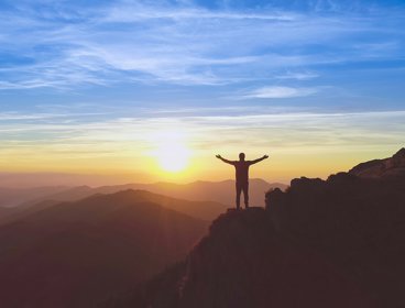 Person standing on a cliff with arms outstretched, in front of a sunset sky