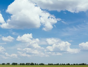 A scene hsowing clouds and blue sky, over a grassy area with trees