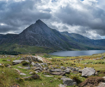 View of grass growth and rocks, a lake and mountain under a cloudy dark sky in Eryri National Park.