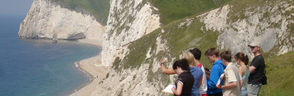 A group of students are sitting near the edge of a cliff, with a view down onto a sandy beach with white chalk cliffs below them.