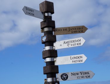 Brown and gray street sign pointing to various cities around the world with distance to them shown too