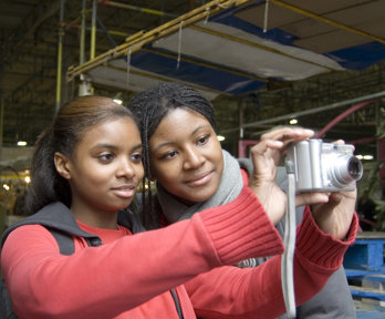 Two female students are in a market area, one is holding a digital camera and taking a picture of the scene. The other is looking at the picture being taken through the camera screen.