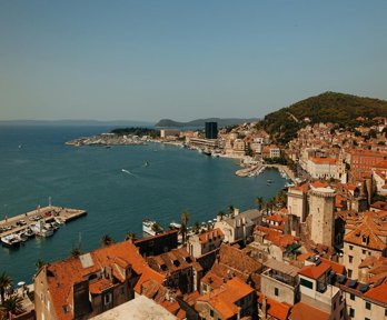 A view over a Mediterranean town by the sea. The roofs are orange tile and several jettys for boats can be seen