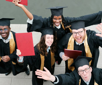 Group of five people in graduation gowns celebrating and raising their certificates.