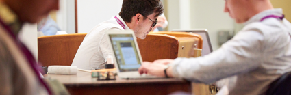 A person in the background but in focus, sits whilst looking downward. Two blurred people in the foreground also sit down, one of which is at a wooden desk typing on a laptop.
