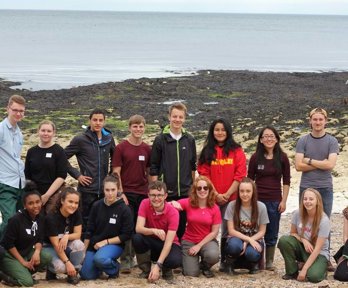 A group of older students lined up on a beach. In the background you can see a rocky beach with seaweed, and then the sea.