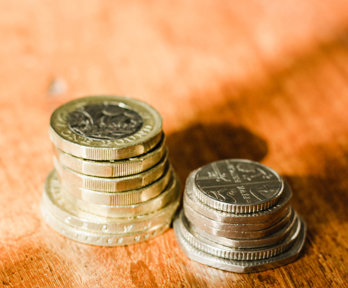 Two piles of english coins, pound coins and two pound coins to the left hand side and fifty pence, twenty pence and five pence to the right hand side, They are placed on a wooden table.