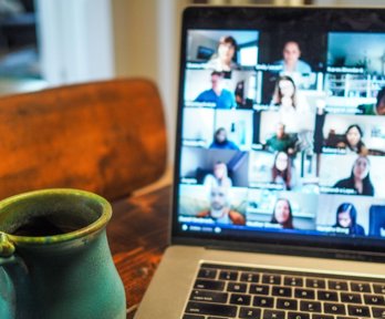 Open laptop showing an online meeting with images of the attendees appearing on the screen. The laptop is sat on a wooden table with a mug next to it.
