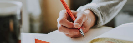 Person holding a pen and writing in an open notebook on a table.