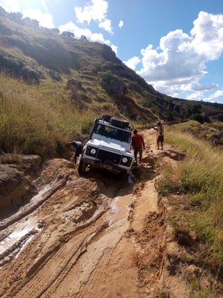 A large car with tyres attached to the roof driving down a mud-covered slope. There are three people stood around the car.