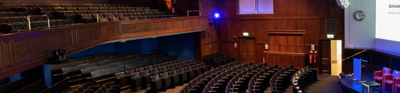 Curved rows of dark brown leather seats occupy the wooden, grey carpeted Royal Geographical Society Ondeetja lecture thearte. The celing is white and the parially visible board is illuminated by the projector.