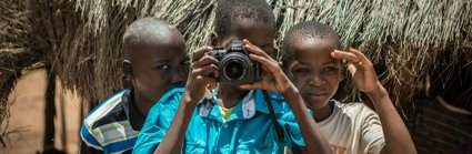 Three African children- one is pointing a camera at the person taking the picture