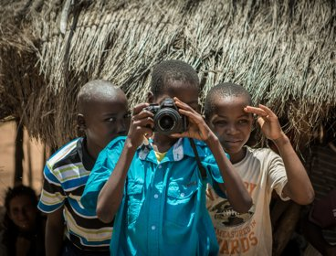 Three African children- one is pointing a camera at the person taking the picture