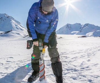 Person using a drill and cylindrical tube to extract an ice core, with snow covered mountains behind.