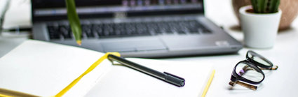 An open notebook with a pen laying on top. A blurred laptop screen in the background shows an ongoing online meeting.