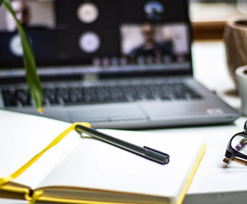 An open notebook with a pen laying on top. A blurred laptop screen in the background shows an ongoing online meeting.