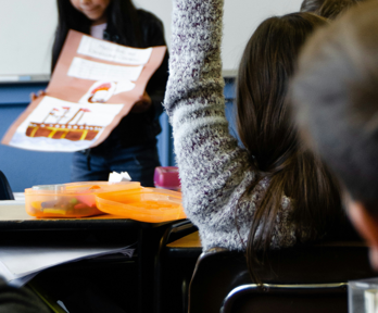 A school child holds their hand up as another child stands in the front of the classroom whilst presenting