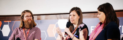 Three people sititng in comfortable chairs on a stage, with the person in the middle holding a microphone and speaking.