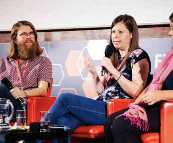 Three people sititng in comfortable chairs on a stage, with the person in the middle holding a microphone and speaking.