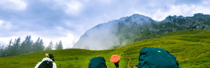 Four people with large backpacks are walking up a green hill towards a rocky outcrop. The clouds are low and are forming a mist at the top of the hill towards where they are heading