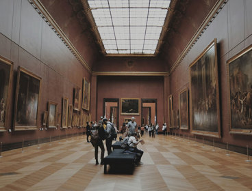 People looking at paintings hung on walls in a gallery hall of Musée du Louvre, Paris, France.