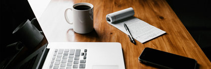 An open laptop, coffee mug containing coffee, notebook, pen and mobile phone on a desk.