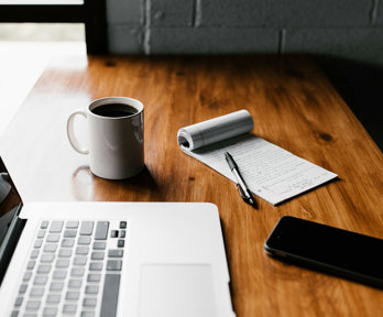 An open laptop, coffee mug containing coffee, notebook, pen and mobile phone on a desk.