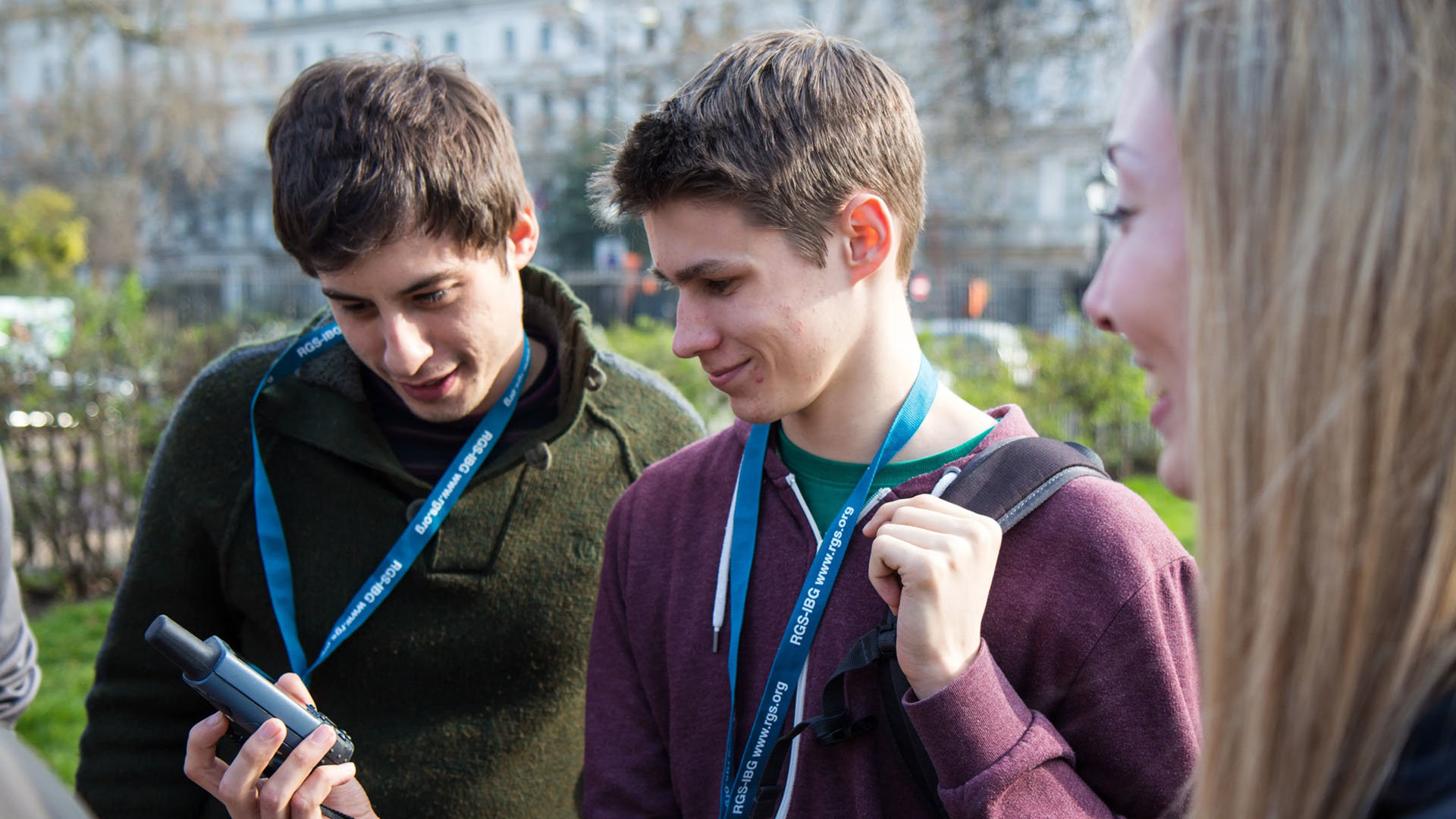 Two young people wearing lanyards look at a measurement device while outside. A third, smiling person watcehs them.