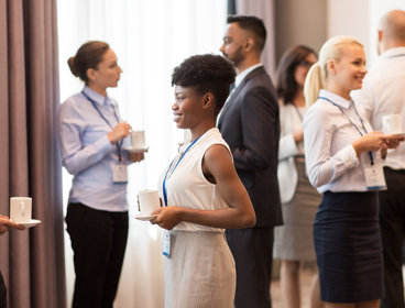 Business people with conference badges and coffee chatting with each other in a brightly lit room.