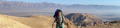 Person in trekking gear standing on top of a hill with a mountain range in the background.