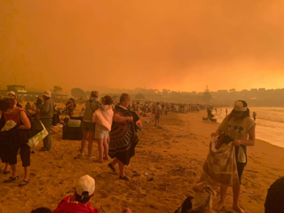 People waiting on a beach to be evacuated. The sky is orange and full of smoke.