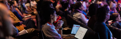 An audience listen to a presentation in a dark lecture hall. 