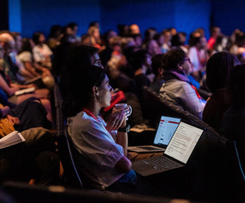 An audience listen to a presentation in a dark lecture hall. 