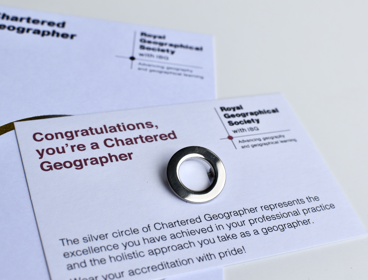 Silver circle pin awarded to Chartered Geographers shown on a white card reading 'Congratulations, you're a Chartered Geographer'