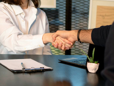 Two people sit across from each other at a table whilst shaking hands. There is a clip board and pen on the table to their left and laptop on the table to their right.