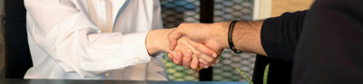 Two people sit across from each other at a table whilst shaking hands. There is a clip board and pen on the table to their left and laptop on the table to their right.