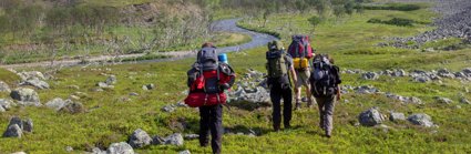 Four people with large rucksacks and camping equipment walking beside a meandering river. There are trees on the bank of the river and piles of rocks across the grass where they are walking.