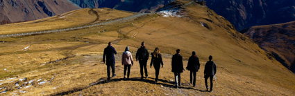 A group of seven people on a brown field facing snow-covered mountains on a sunny day.
