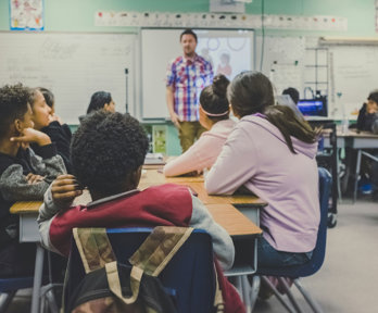 A teacher standing at the front of a classroom with students sat at the desks.