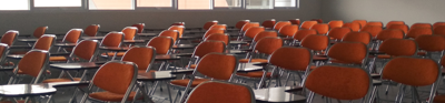 Orange chairs neatly fill an empty class room. large square panel windows on the left of the room let in beams of light.